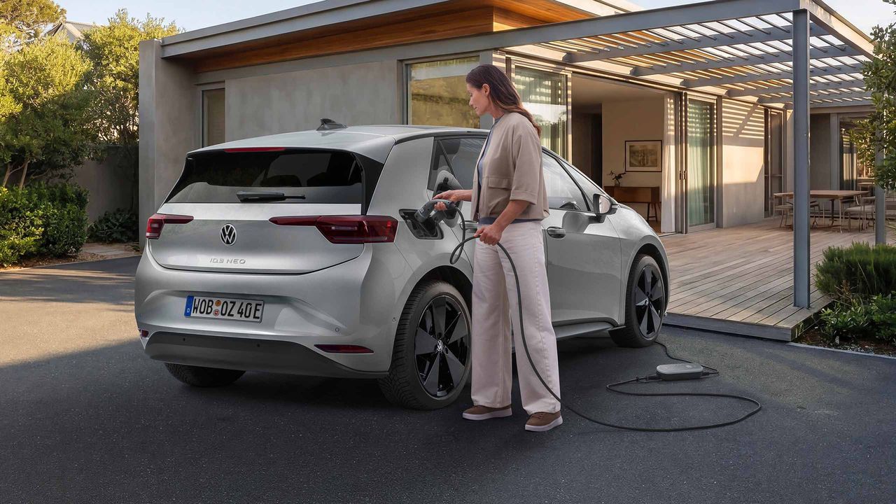 Млада жена зарежда сив електрически автомобил Volkswagen, паркиран пред модерна къща с големи стъклени прозорци. Тя държи кабела за зареждане, включен в автомобила, а зарядното устройство е на земята до нея. На заден план се вижда подреден двор с зеленина и частично покрито пространство с дървена конструкция.