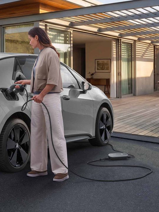 Млада жена зарежда сив електрически автомобил Volkswagen, паркиран пред модерна къща с големи стъклени прозорци. Тя държи кабела за зареждане, включен в автомобила, а зарядното устройство е на земята до нея. На заден план се вижда подреден двор с зеленина и частично покрито пространство с дървена конструкция.