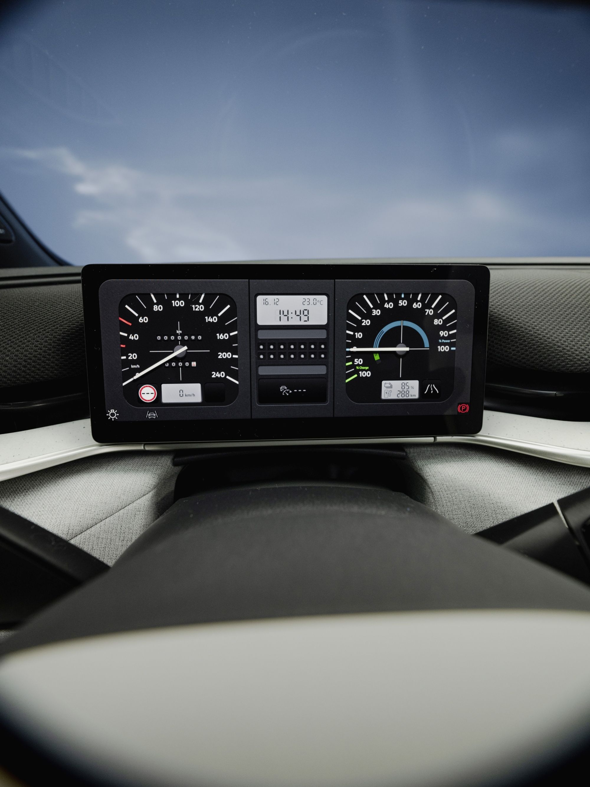 Изображението показва приборното табло на автомобил с два аналогови скоростомера и цифров дисплей, показващ час. Таблото е модерно и има различни индикатори за състоянието на автомобила. Небето се вижда през предното стъкло.