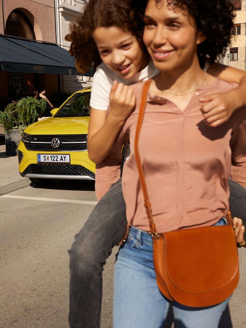 На централна градска улица жена носи на гърба си момиче с жълт VW T-Cross на заден план.