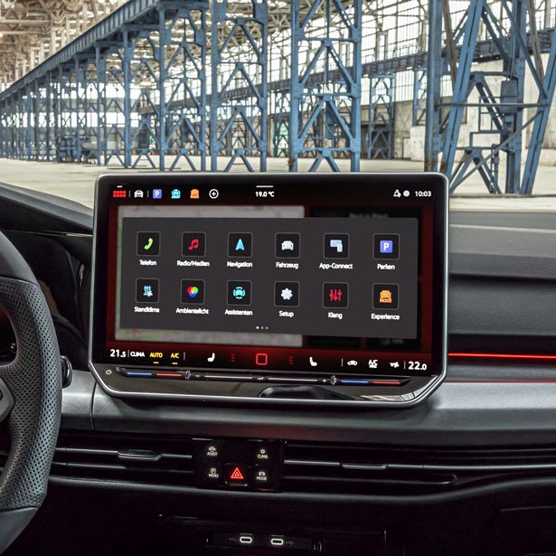 Das Bild zeigt das Cockpit eines modernen Golf GTI. Auffällig ist das große Touchscreen-Bedienfeld und die digitale Instrumententafel. Im Hintergrund ist eine große Industriehalle zu sehen.
