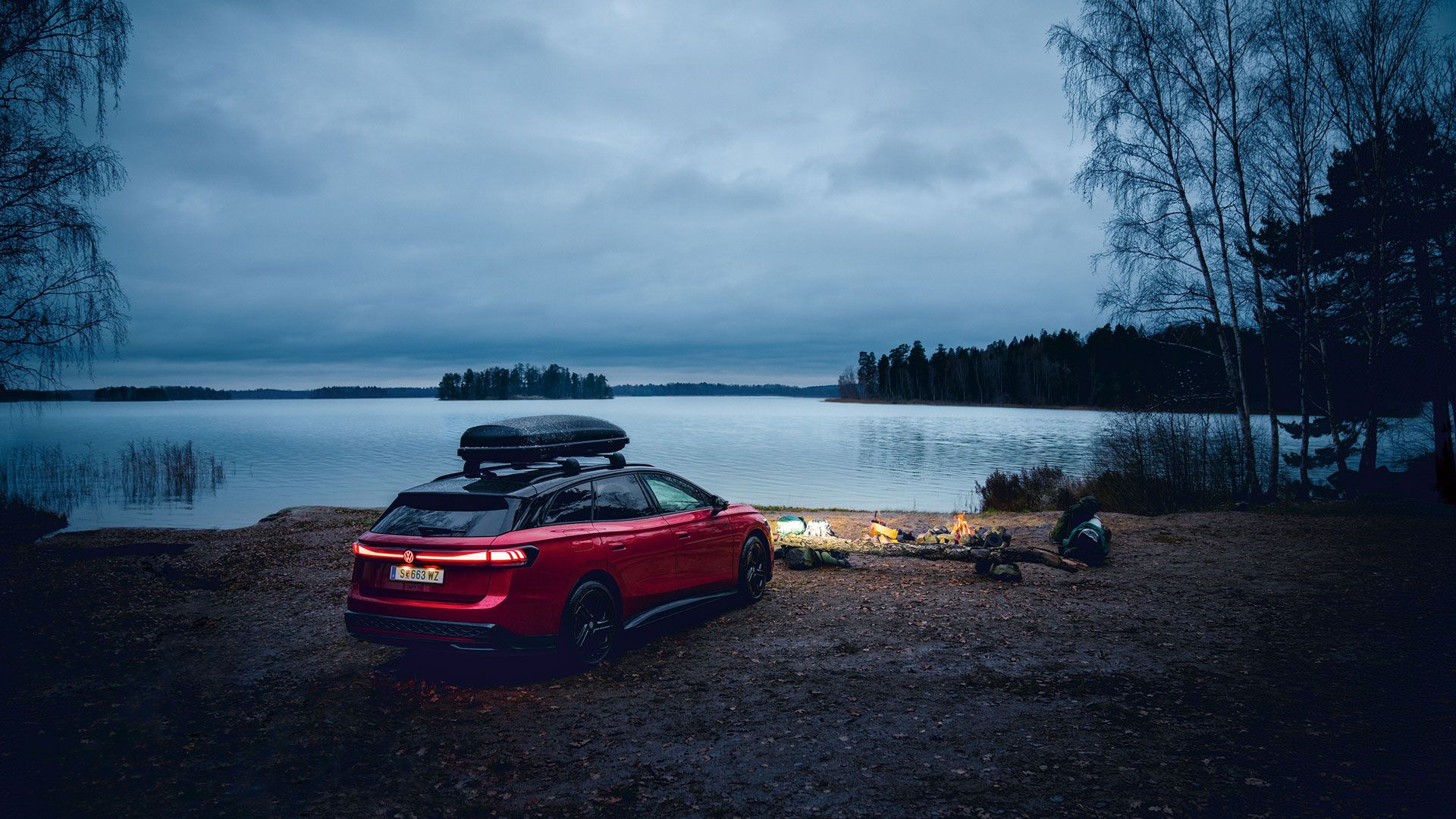 Червен Volkswagen ID.7 Tourer с покривен багажник, паркиран край езеро до вечерен лагерен огън