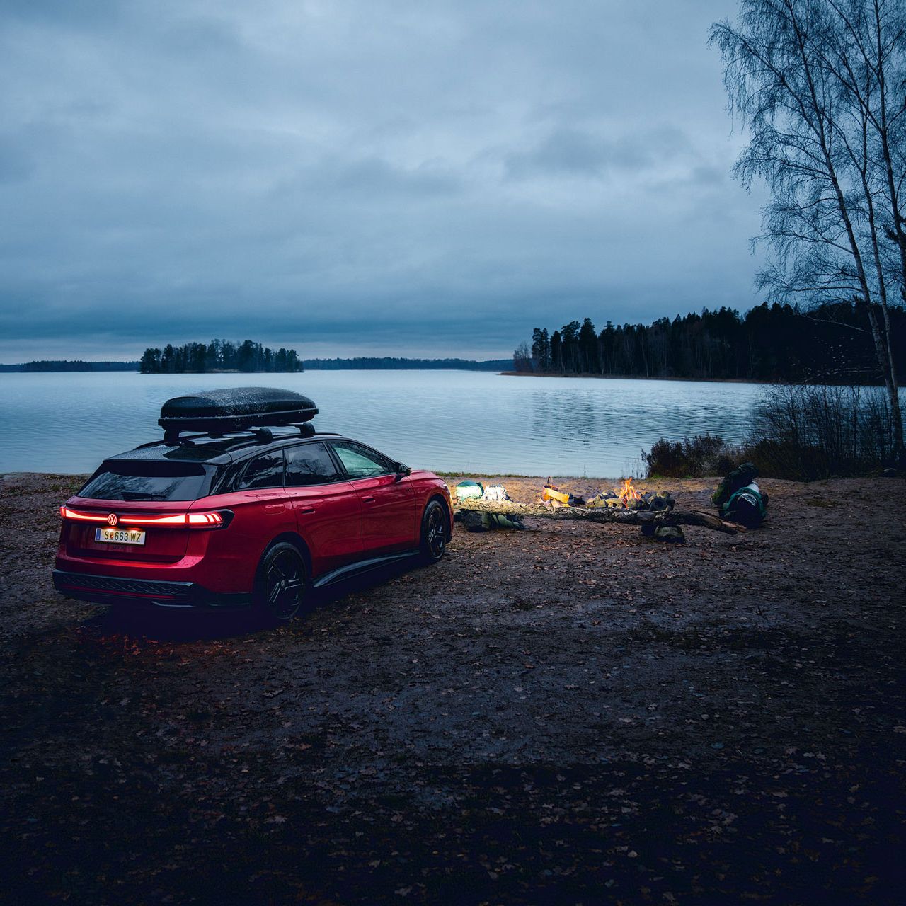 Червен Volkswagen ID.7 Tourer с покривен багажник, паркиран край езеро до вечерен лагерен огън