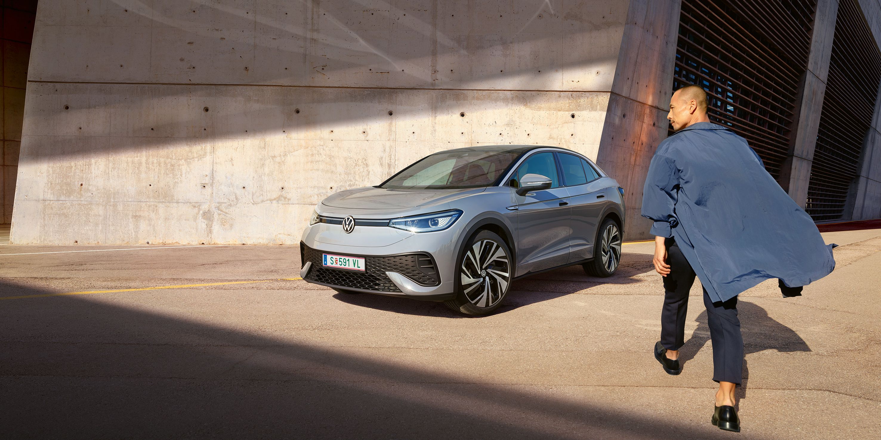 Grauer VW ID.5 parkt vor heller Hauswand, Ansicht schräg rechts von vorn, Mann geht zügig auf das E-Auto zu.