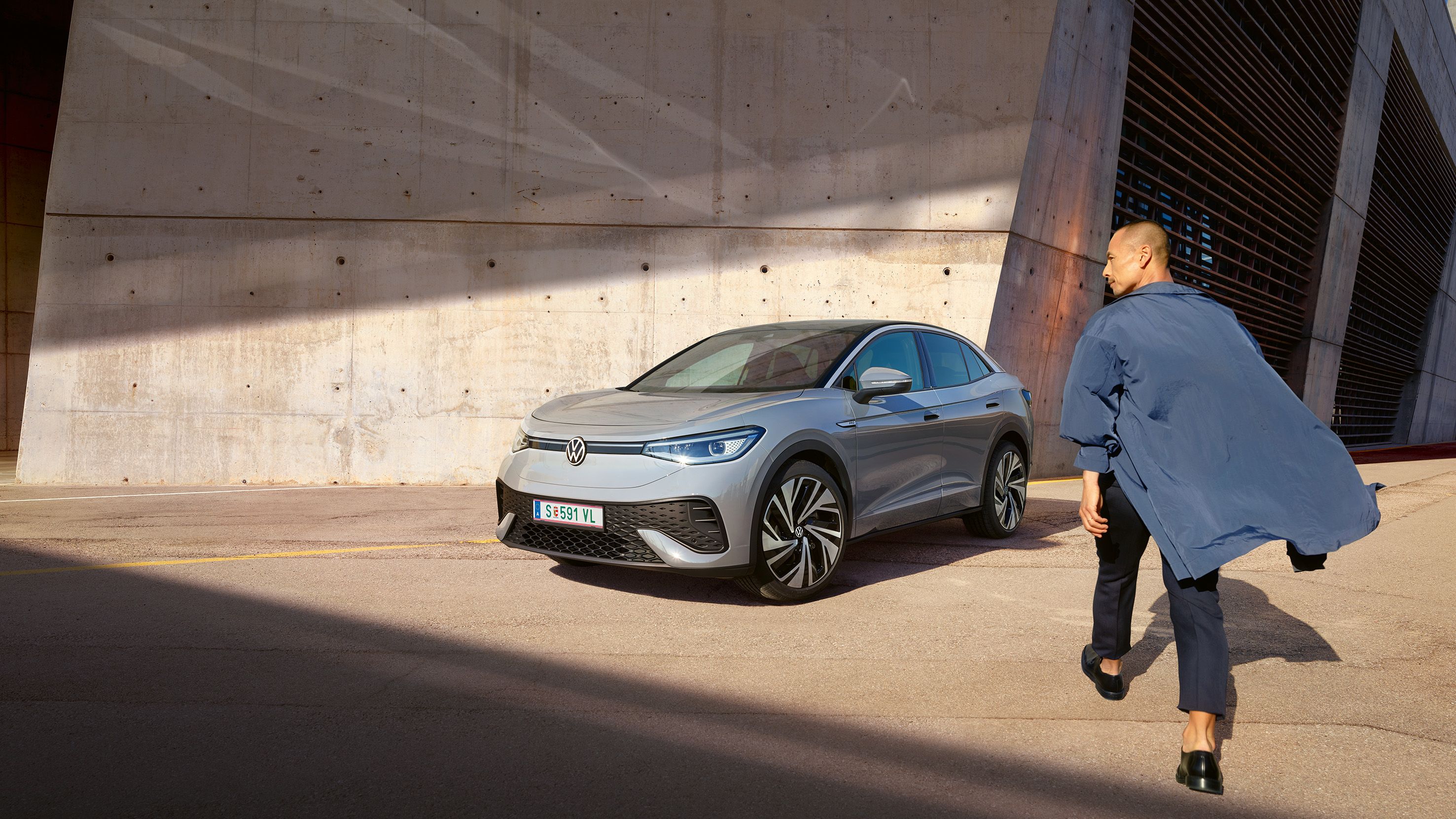 Grauer VW ID.5 parkt vor heller Hauswand, Ansicht schräg rechts von vorn, Mann geht zügig auf das E-Auto zu.