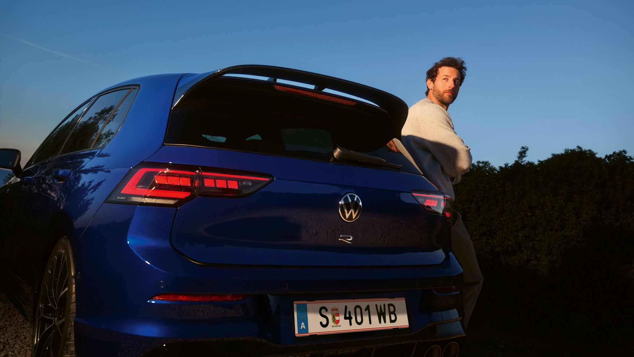 Mann lehnt sich seitlich an den Golf R an, welcher in Heckansicht steht.