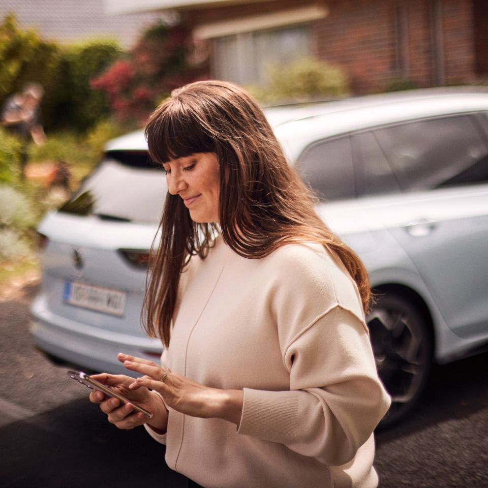 Eine junge Frau steht hinter einem weißen VW Golf Estate und benutzt ihr Smartphone.