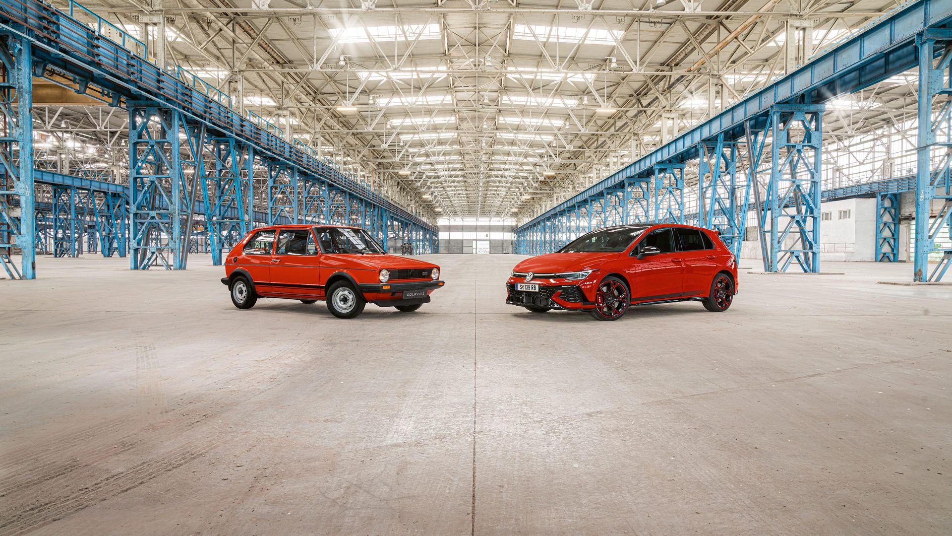 Zwei rote Golf GTI, das erste und das neuste Modell, stehen in einer großen Industriehalle mit vielen blauen Stahlträgern und einem hohen, hellen Dach.