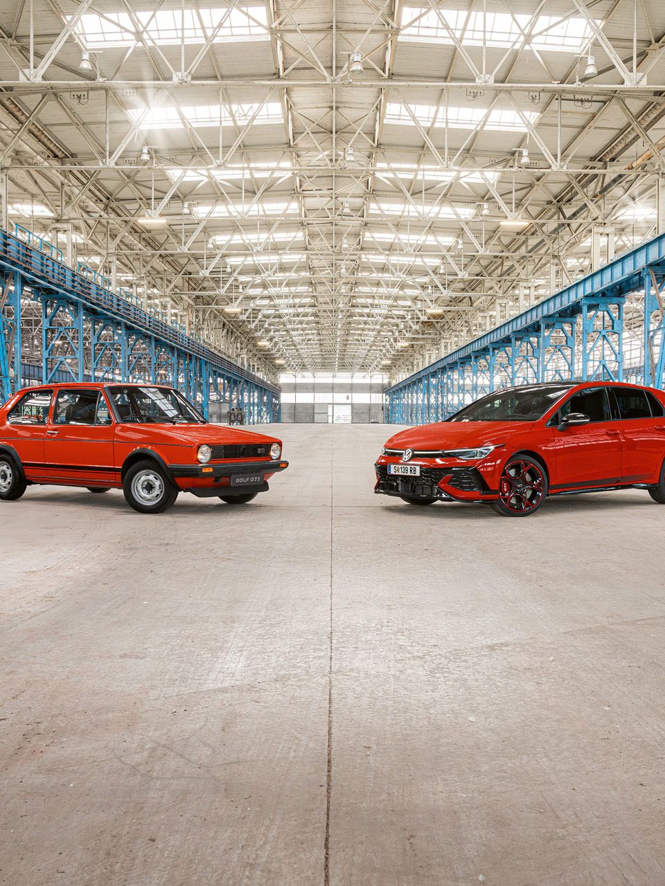 Zwei rote Golf GTI, das erste und das neuste Modell, stehen in einer großen Industriehalle mit vielen blauen Stahlträgern und einem hohen, hellen Dach.