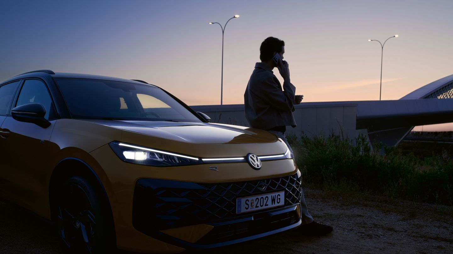 Жълт Volkswagen T-Roc R-Line в здрач с включени предни светлини и силует на мъж до автомобила.
