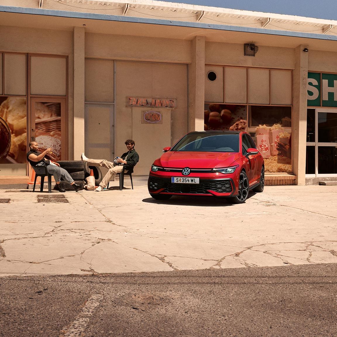 Червен Volkswagen Golf пред малък магазин с хора, които си почиват на сянка