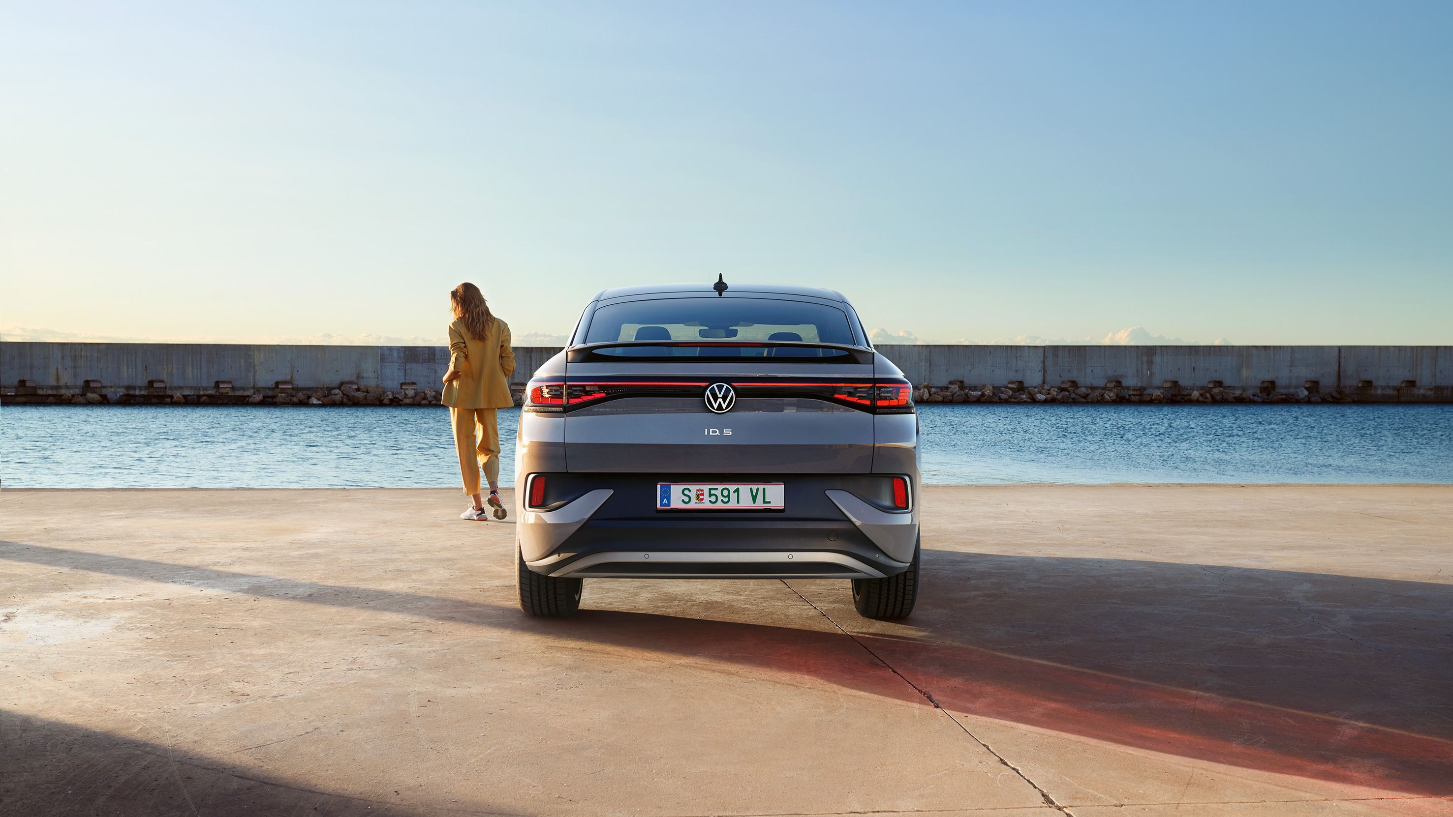 Heckansicht eines VW ID.5 an einem Hafenkai, neben dem E-Auto steht eine Frau