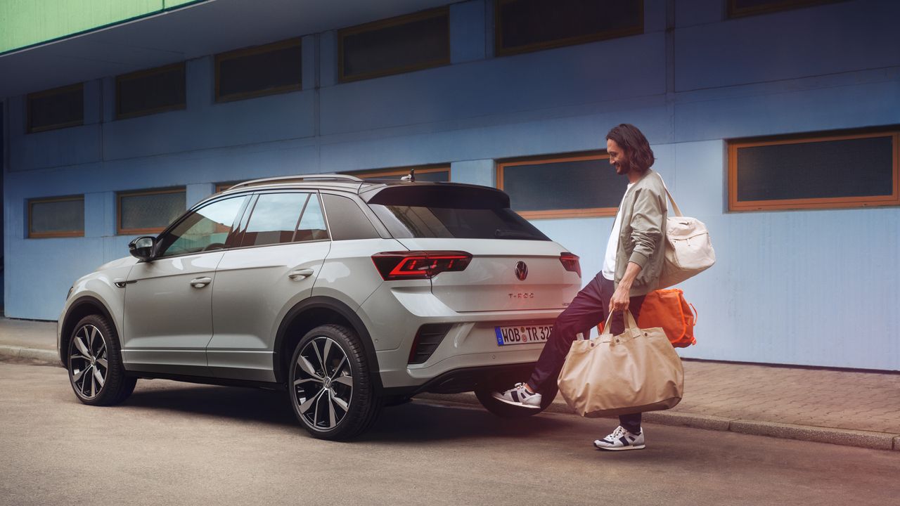 VW T-Roc R-Line in weiß vor einem Gebäude, Seite sichtbar, Mann steht hinter dem Auto und öffnet mit Fußschwenk die Kofferraumklappe
