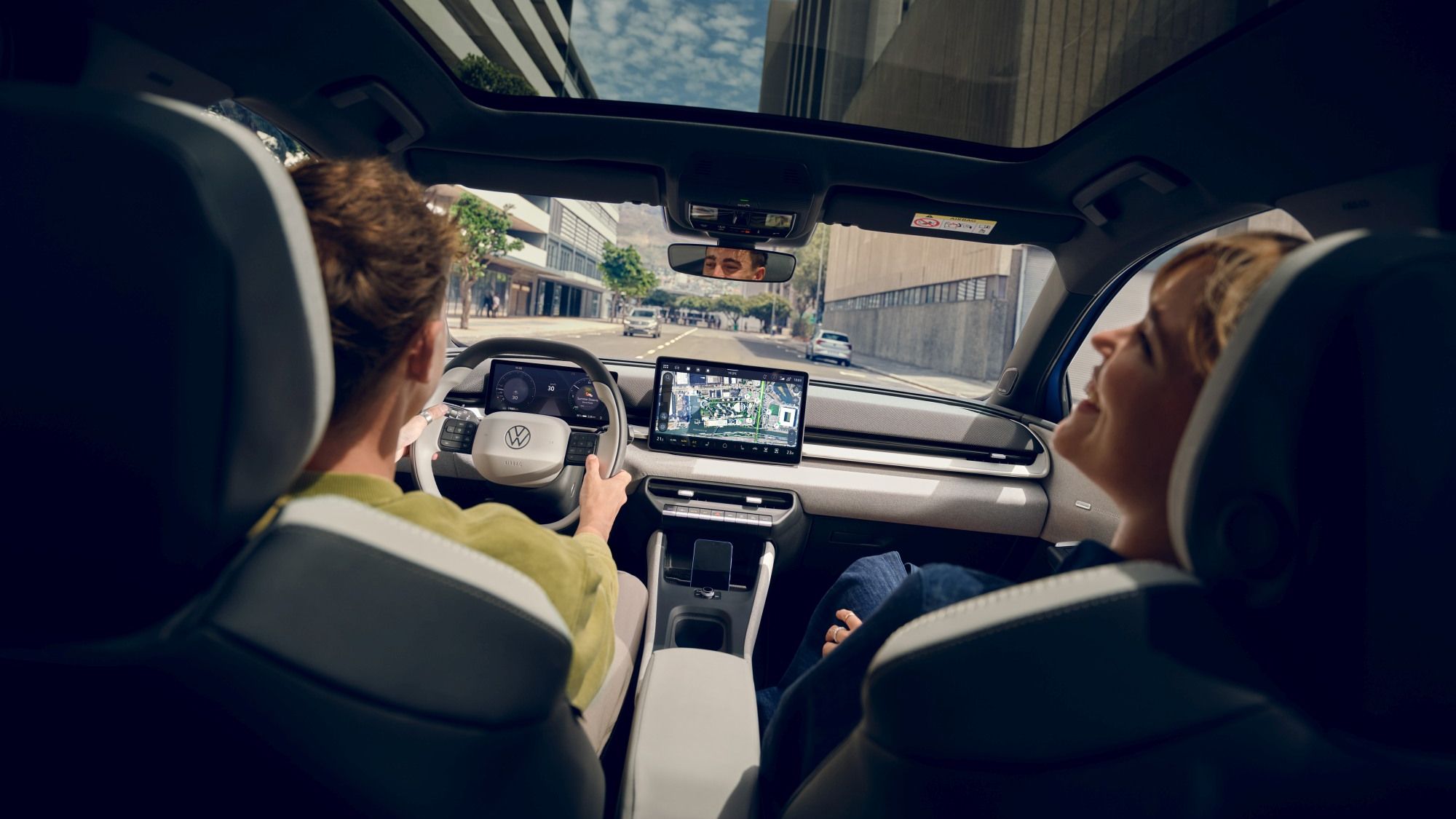 Изглед отзад към двама души на предните седалки на Volkswagen ID.Polo с панорамен стъклен покрив. Шофьорът държи волана и гледа напред, а до него пътникът се е облегнал назад и се наслаждава на гледката. През големия покрив се виждат високи офис сгради и синьо небе с облаци.