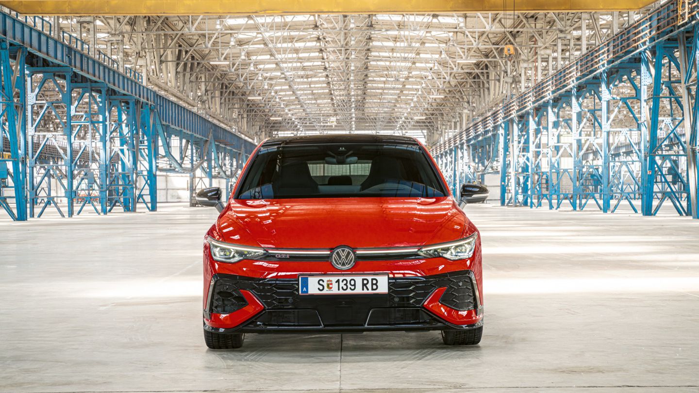 Ein roter Golf GTI steht in einer großen, hell beleuchteten Lagerhalle. Das Auto ist von vorne zu sehen und hat das Kennzeichen MÖB-GT 150. Im Hintergrund sind metallene Strukturen und Säulen zu sehen.