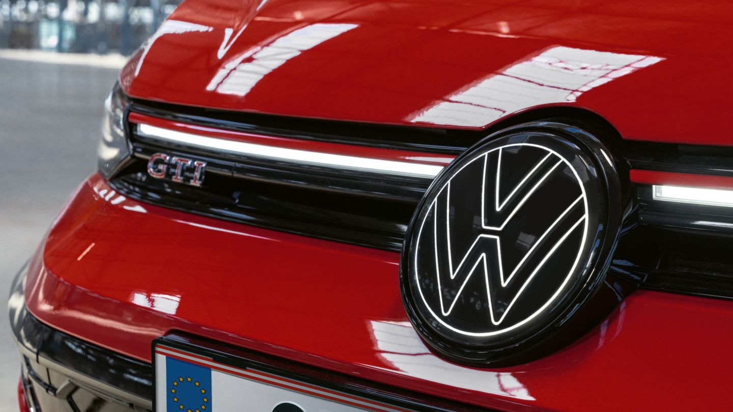 Nahaufnahme der Front eines roten Golf GTI mit GTI Schriftzug und dem markanten VW-Logo. Ein EU-Kennzeichen ist ebenfalls sichtbar. Das Auto steht in einer mittelgroßen Garage oder Halle.
