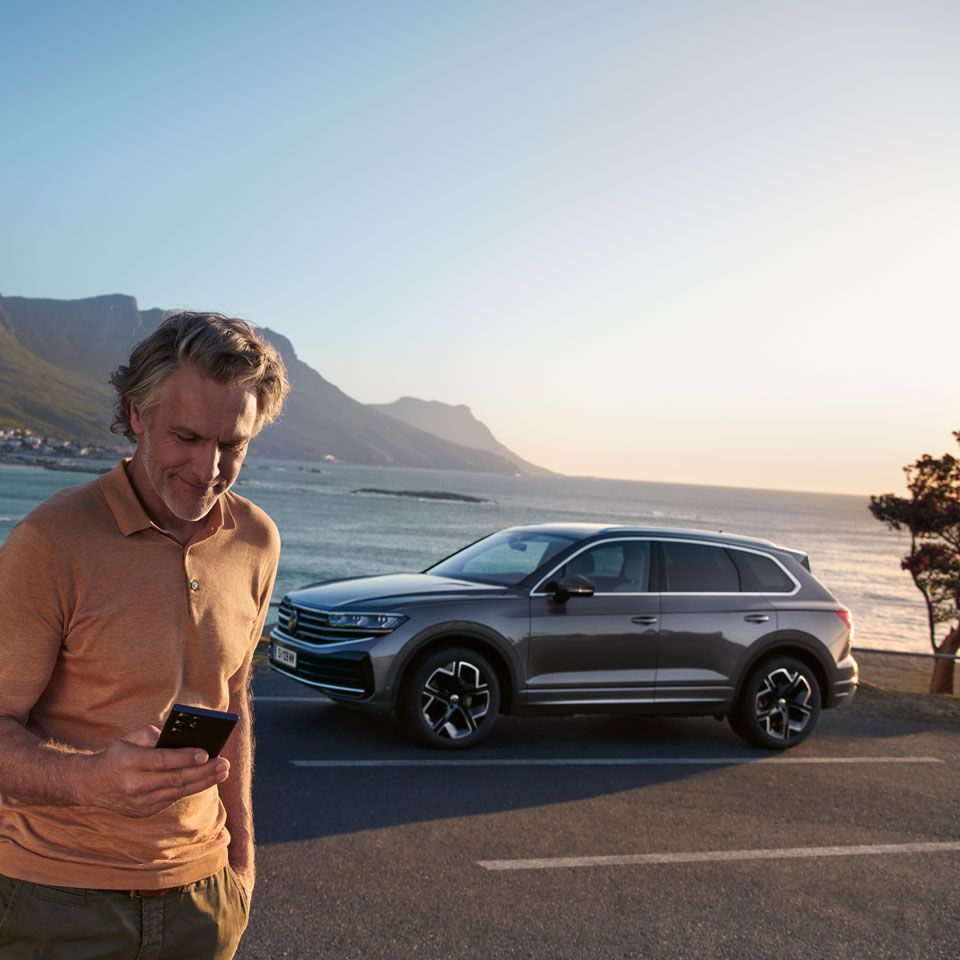 Мъж гледа в смартфона си, зад него е паркиран VW Touareg Elegance, а на заден план е морето и брега.