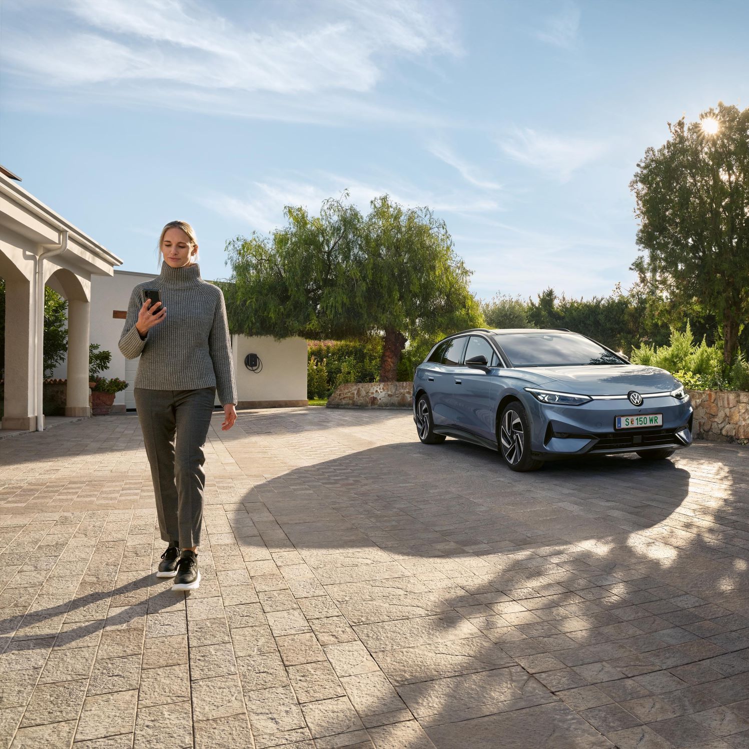Жена с мобилен телефон пред паркиран Volkswagen ID.7 Tourer в двор с дървета