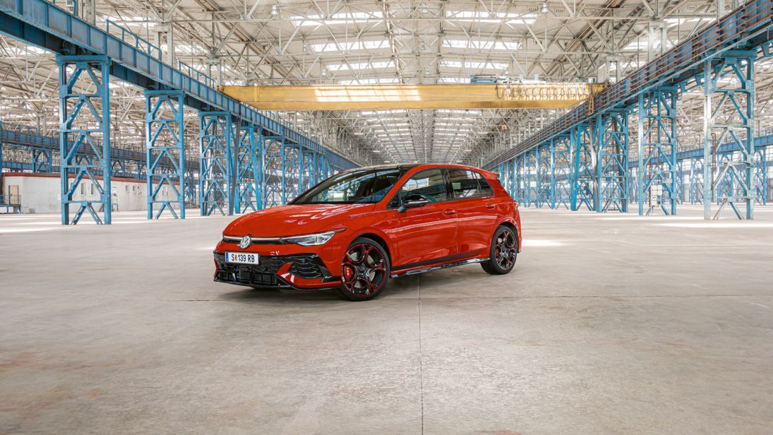 Ein roter Golf GTI steht in einer großen, leeren Industriehalle mit blauen Stahlträgern. Das Dach der Halle ist aus Glas und lässt viel Licht herein.