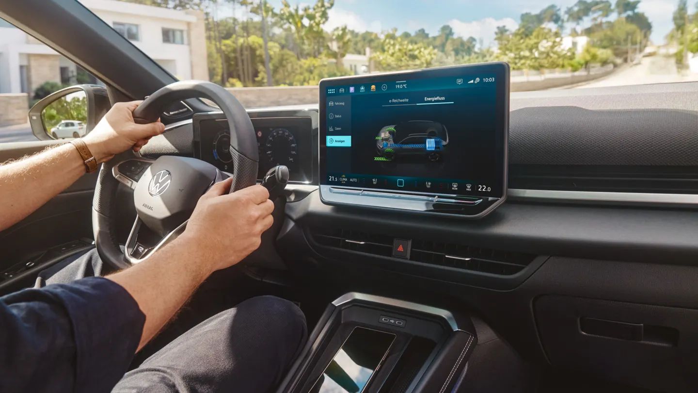 Човек шофира модерен автомобил Volkswagen T-Roc Full Hybrid, като държи волана с две ръце. На големия централен дисплей е показан интерфейс на мултимедийната и навигационната система с визуализация на автомобила. През предното стъкло се вижда слънчева улица с къщи и дървета.