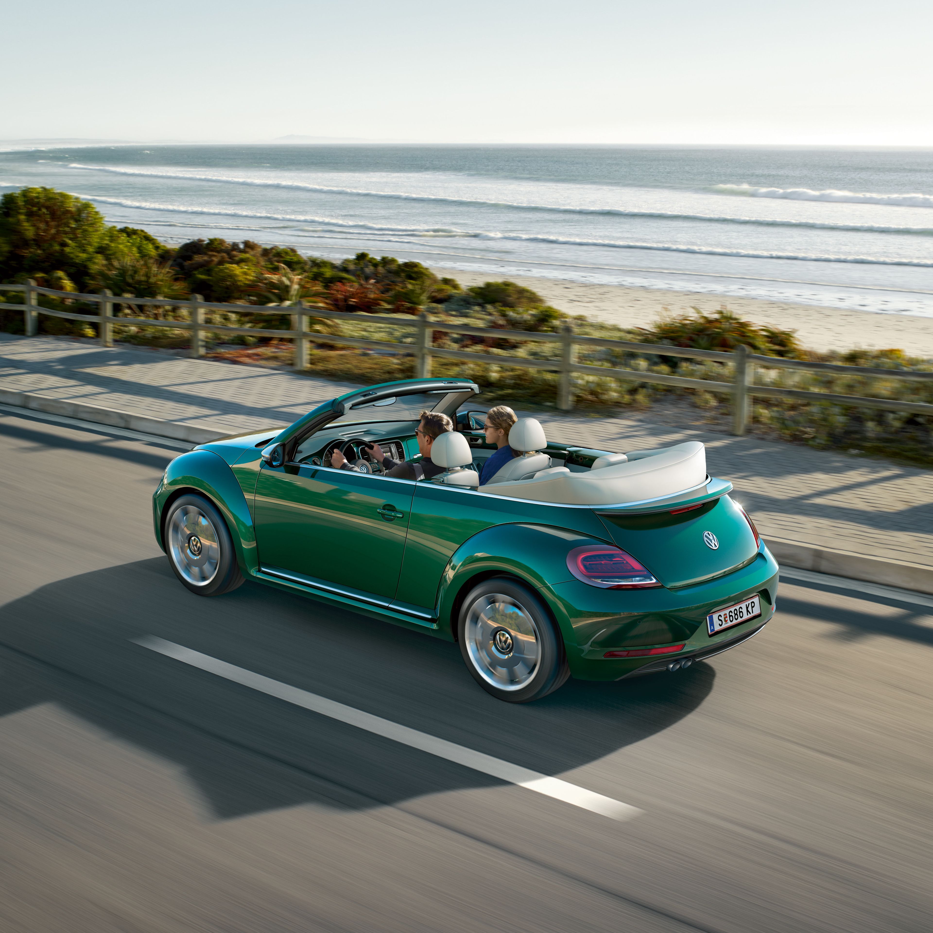 Зелен Volkswagen Beetle кабриолет в движение по крайбрежен път с отворен покрив.