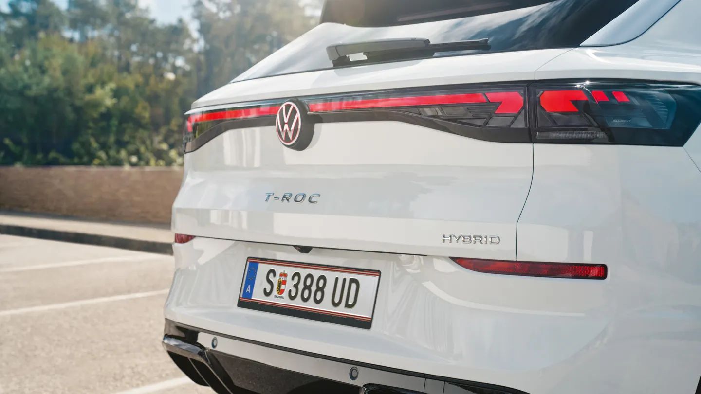 Задна част на бял Volkswagen T-Roc Full Hybrid, паркиран на открито. На снимката ясно се виждат емблемата на VW, моделното обозначение T-ROC, надписът Hybrid и европейски регистрационен номер. В далечината се забелязват дървета и ниска стена.