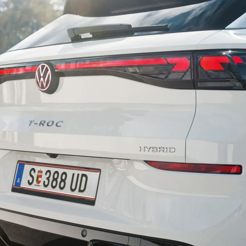 Задна част на бял Volkswagen T-Roc Full Hybrid, паркиран на открито. На снимката ясно се виждат емблемата на VW, моделното обозначение T-ROC, надписът Hybrid и европейски регистрационен номер. В далечината се забелязват дървета и ниска стена.