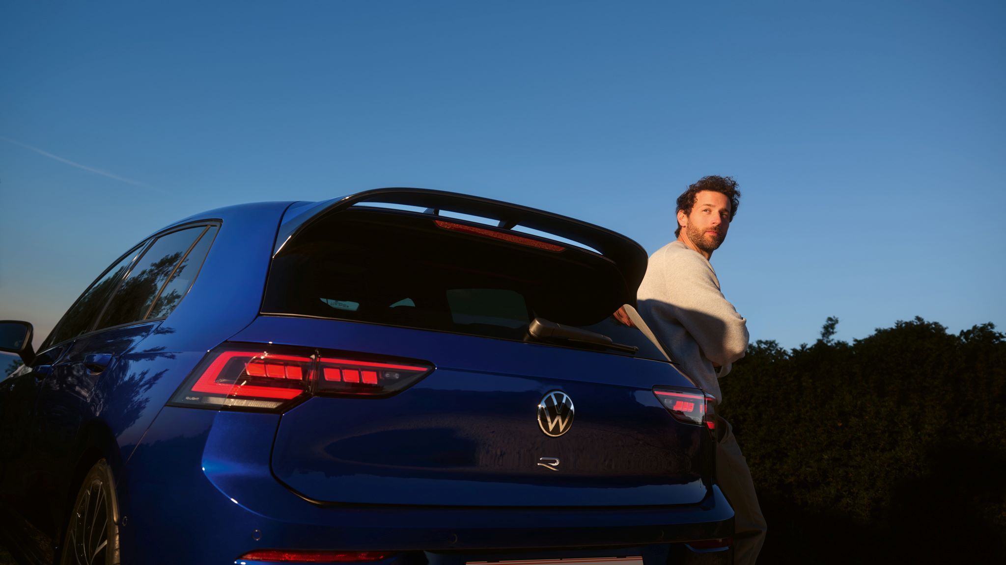 Mann lehnt sich seitlich an den Golf R an, welcher in Heckansicht steht.