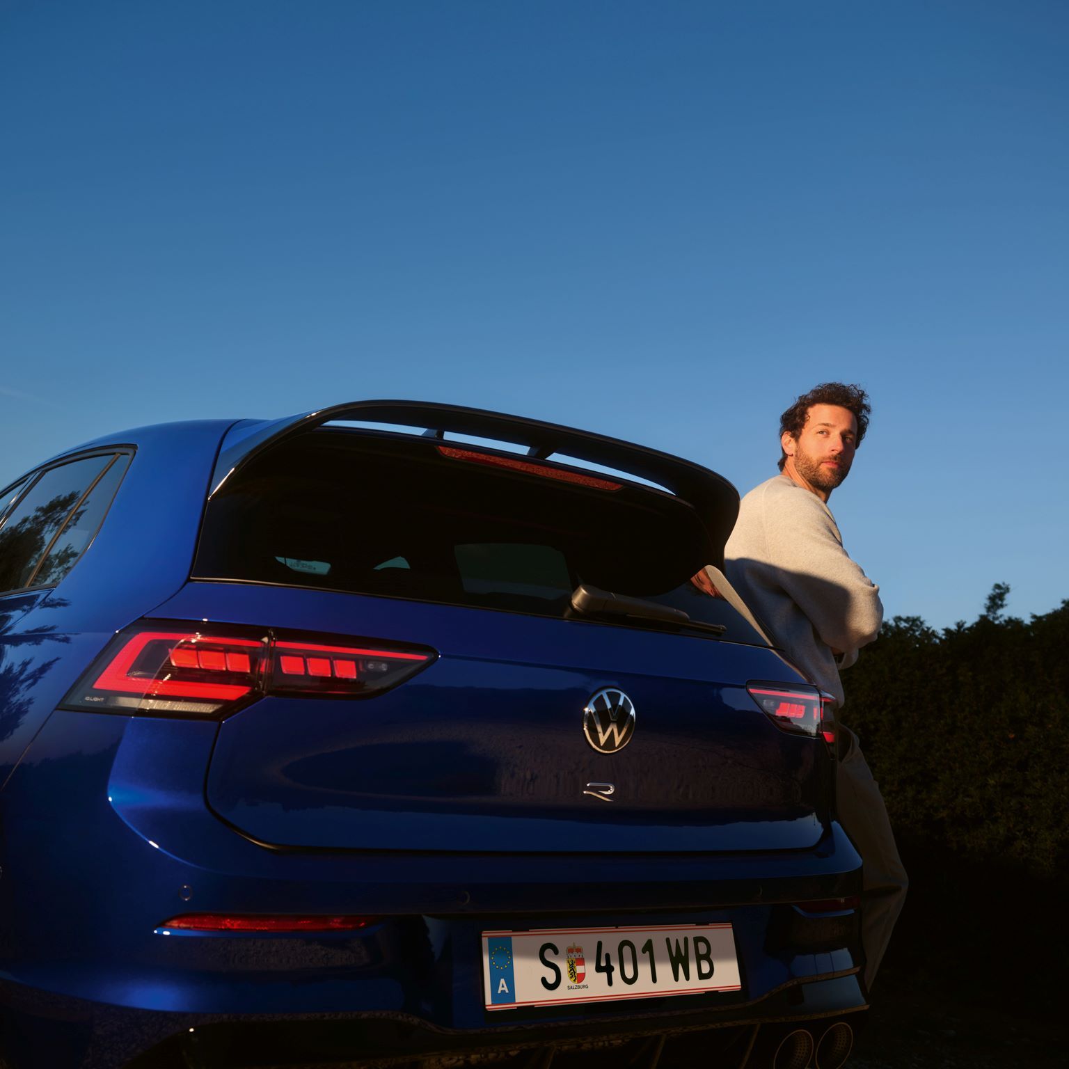 Mann lehnt sich seitlich an den Golf R an, welcher in Heckansicht steht.