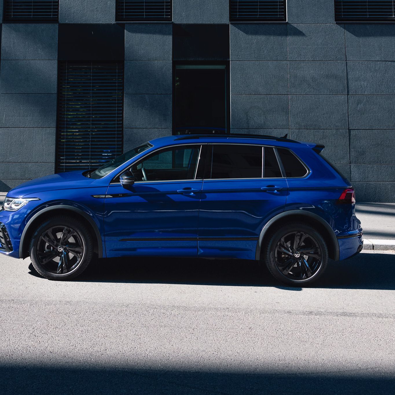 Син Volkswagen Tiguan R, заснет в профил, паркиран на улица до фасада на съвременна офис сграда