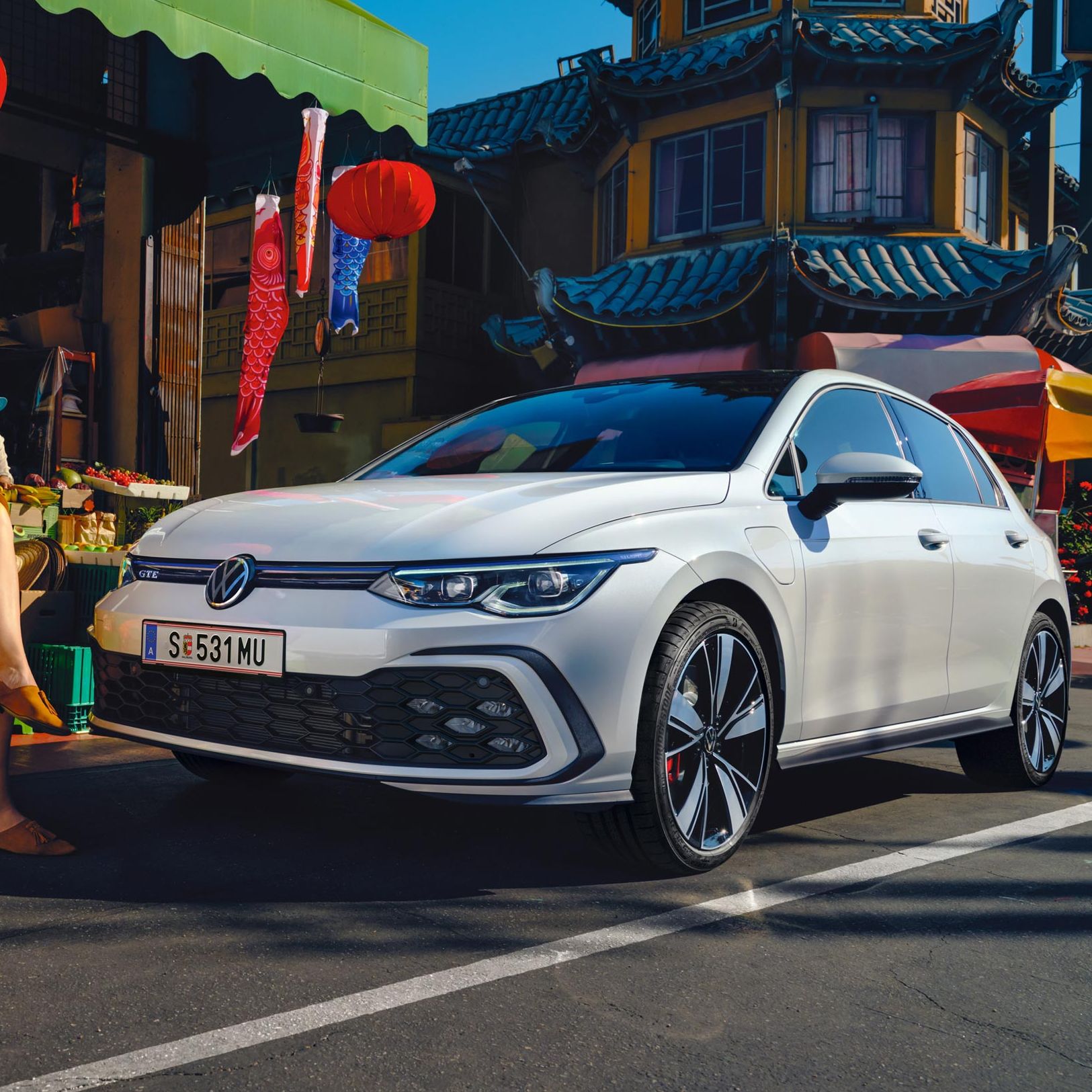 Бял Volkswagen Golf GTE паркиран пред пазар с хора и цветни декорации в оживена градска среда