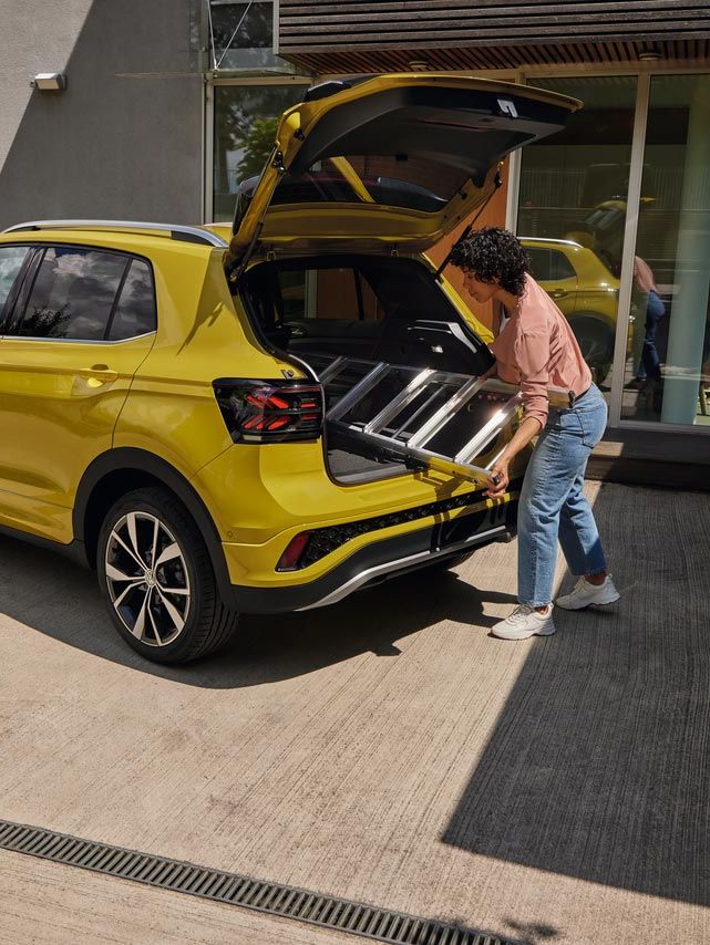 Eine junge Frau schiebt eine Leiter in den Kofferraum eines gelben VW T-Cross.