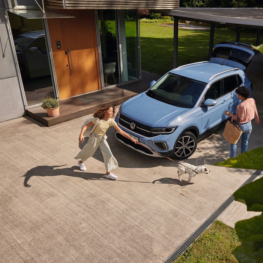 Жена с кошница и момиче с куче пред син VW T-Cross с отворен багажник, паркиран на алея.
