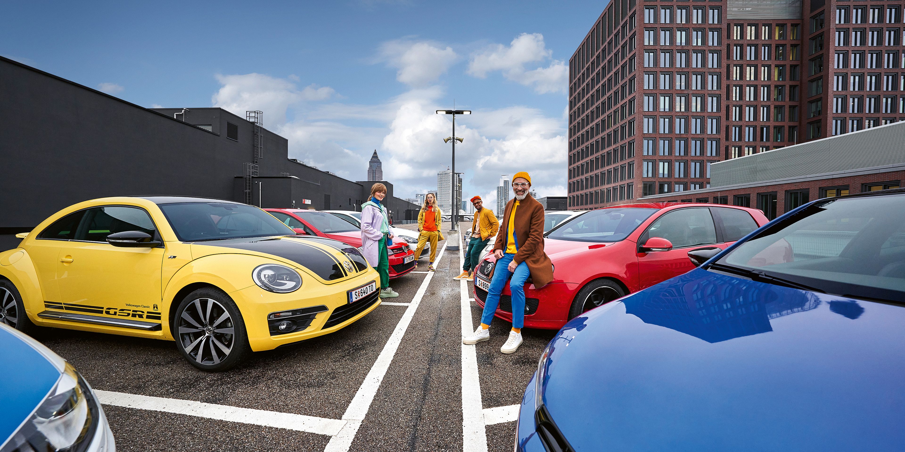 Малка групичка от хора, заобиколени от различни VW модели, са се облегнали на тях. Жълт Beetle, Golf GTI различни моделни генерации и други. В даличината се виждат бизнес сгради.