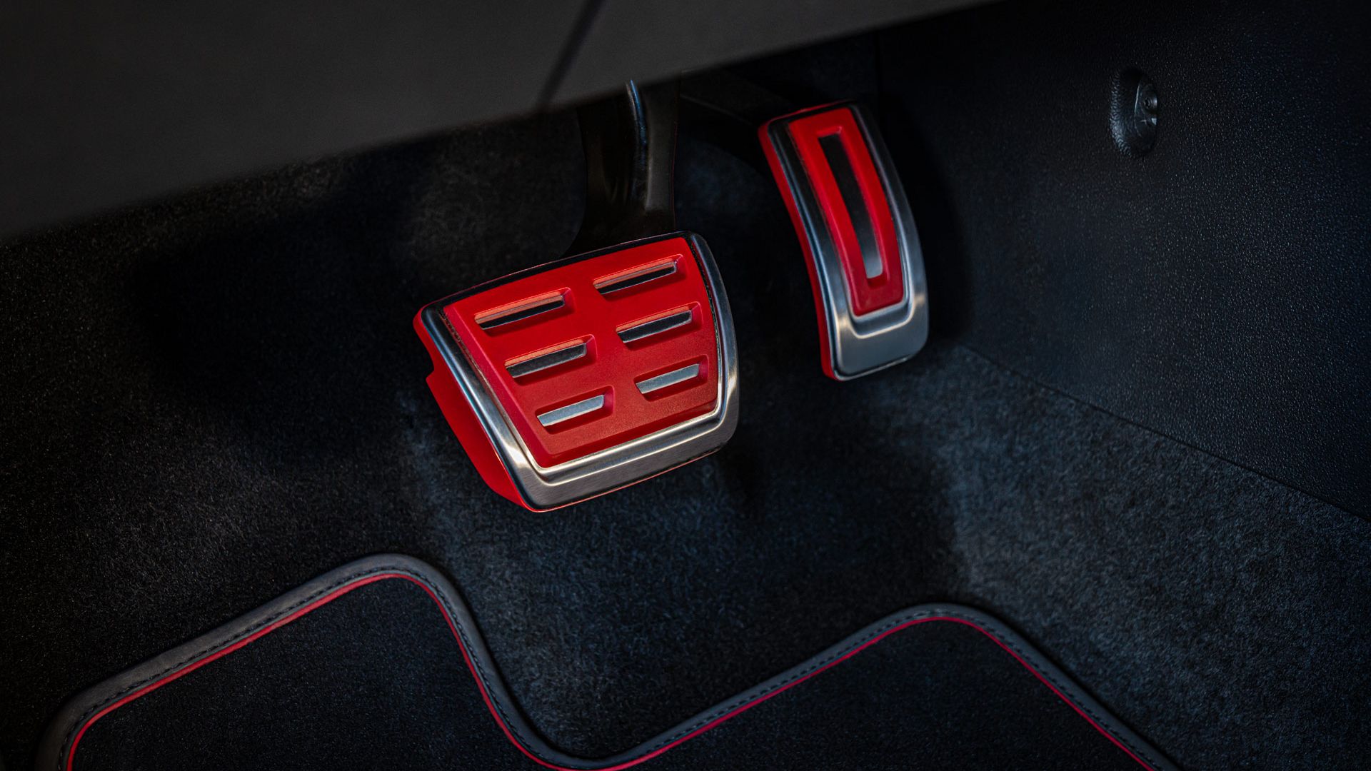 Nahaufnahme der Pedale eines Autos mit schwarzem Teppichboden. Die Pedale haben rote und silberne Akzente. Links ist das Bremspedal und rechts das Gaspedal.