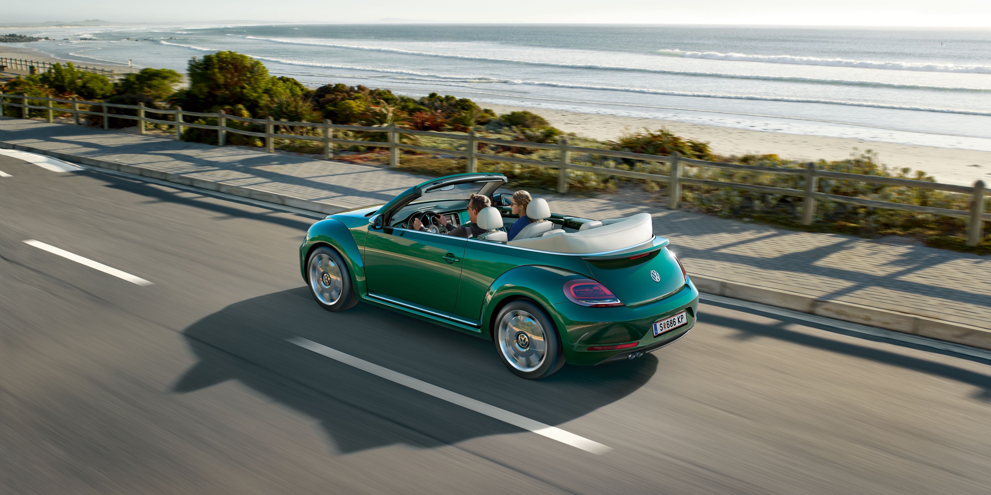 Зелен Volkswagen Beetle кабриолет в движение по крайбрежен път с отворен покрив.