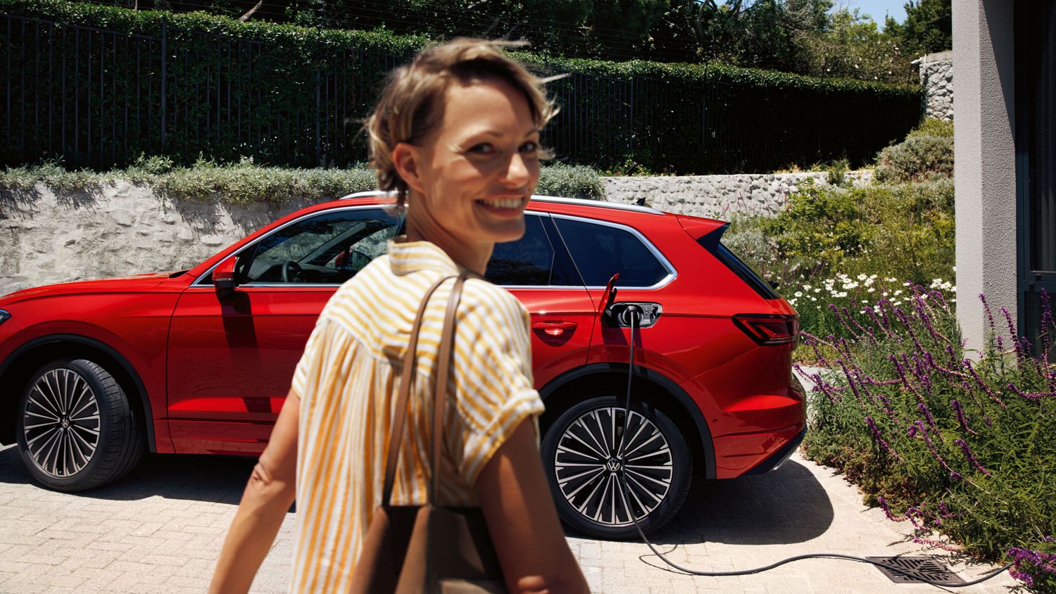 Усмихната жена на преден план, VW Touareg Elegance eHybrid зарежда на зарядна станция на заден план.