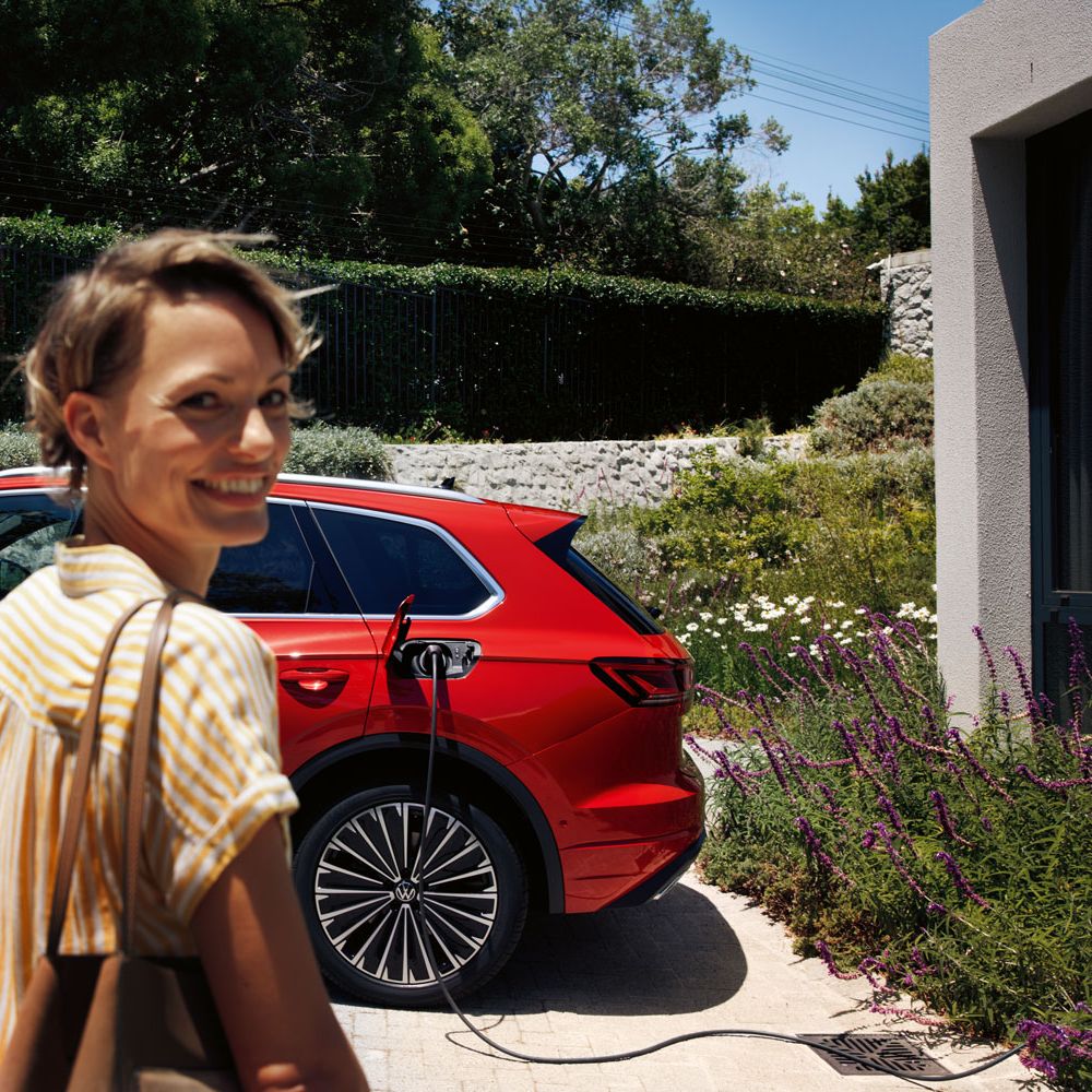 Усмихната жена на преден план, VW Touareg Elegance eHybrid зарежда на зарядна станция на заден план.