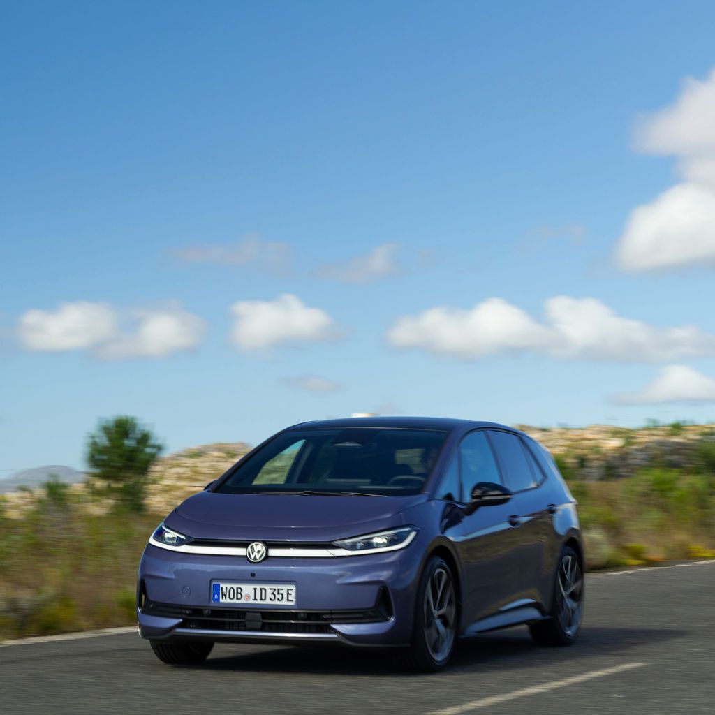 Син електрически хечбек Volkswagen ID.3 се движи по пуст междуградски път при ясно време. Наоколо има ниска растителност и хълмист пейзаж под синьо небе с разпръснати облаци. Колата е заснета в движение, леко отляво отпред.