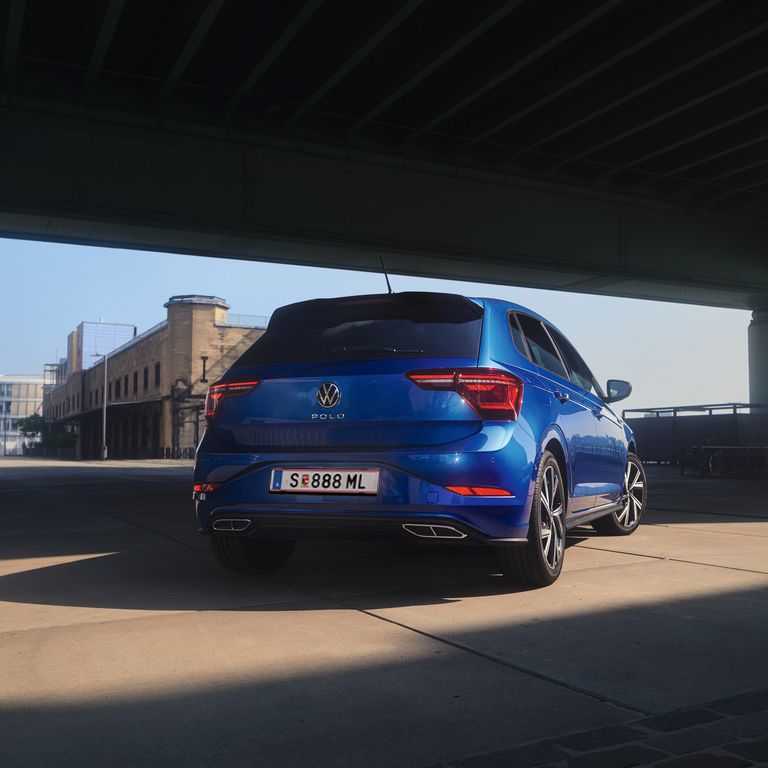 Синьо Volkswagen Polo, заснето отзад под мост в градска среда.