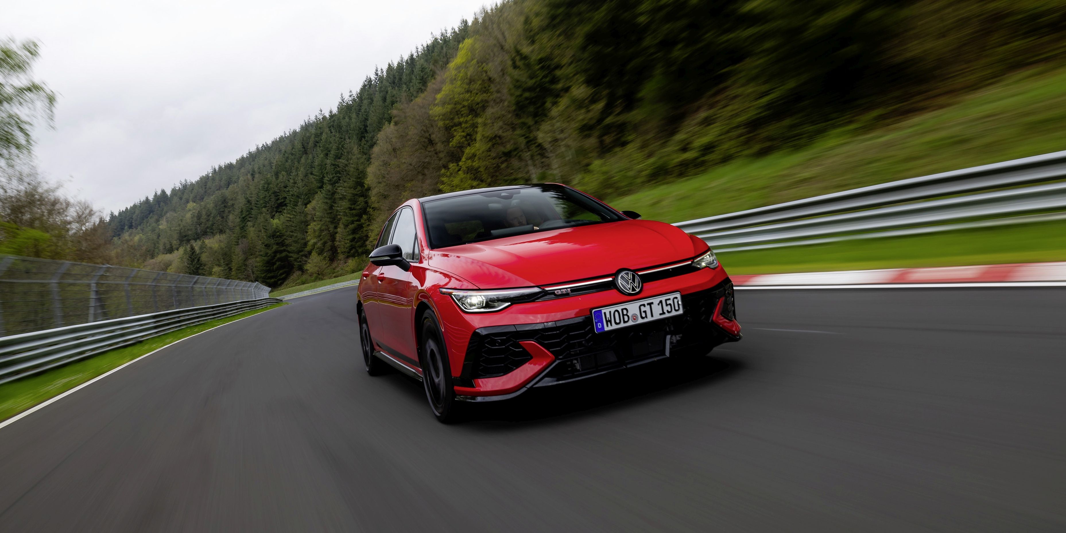 Червен Golf GTI се движи по състезателна писта, заобиколена от зелени дървета и метална ограда. На предната част на автомобила има регистрационен номер. Автомобилът е заснет в движение.