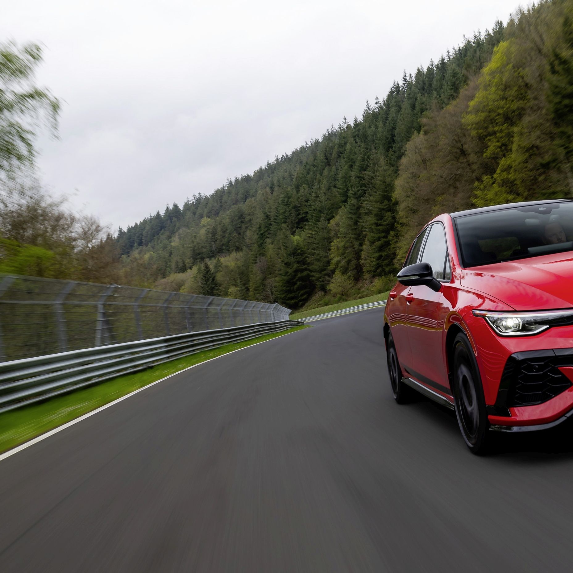 Червен Golf GTI се движи по състезателна писта, заобиколена от зелени дървета и метална ограда. На предната част на автомобила има регистрационен номер. Автомобилът е заснет в движение.