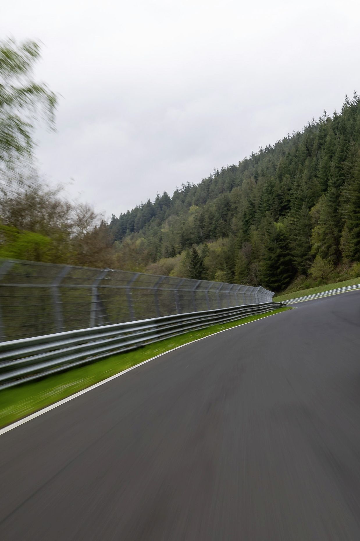 Червен Golf GTI се движи по състезателна писта, заобиколена от зелени дървета и метална ограда. На предната част на автомобила има регистрационен номер. Автомобилът е заснет в движение.