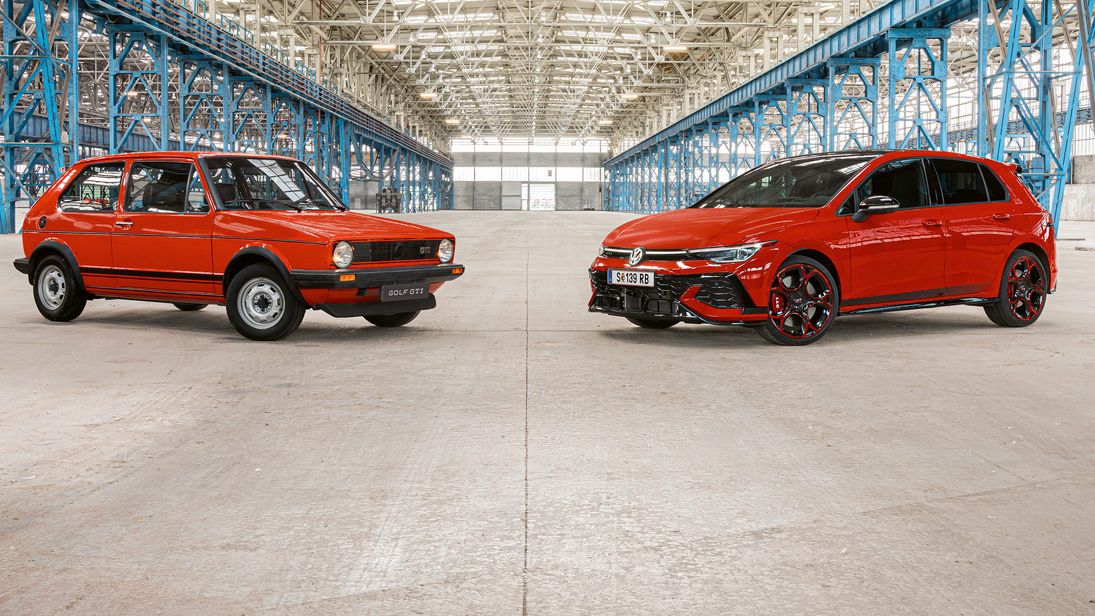 Zwei rote Golf GTI, das erste und das neuste Modell, stehen in einer großen Industriehalle mit vielen blauen Stahlträgern und einem hohen, hellen Dach.