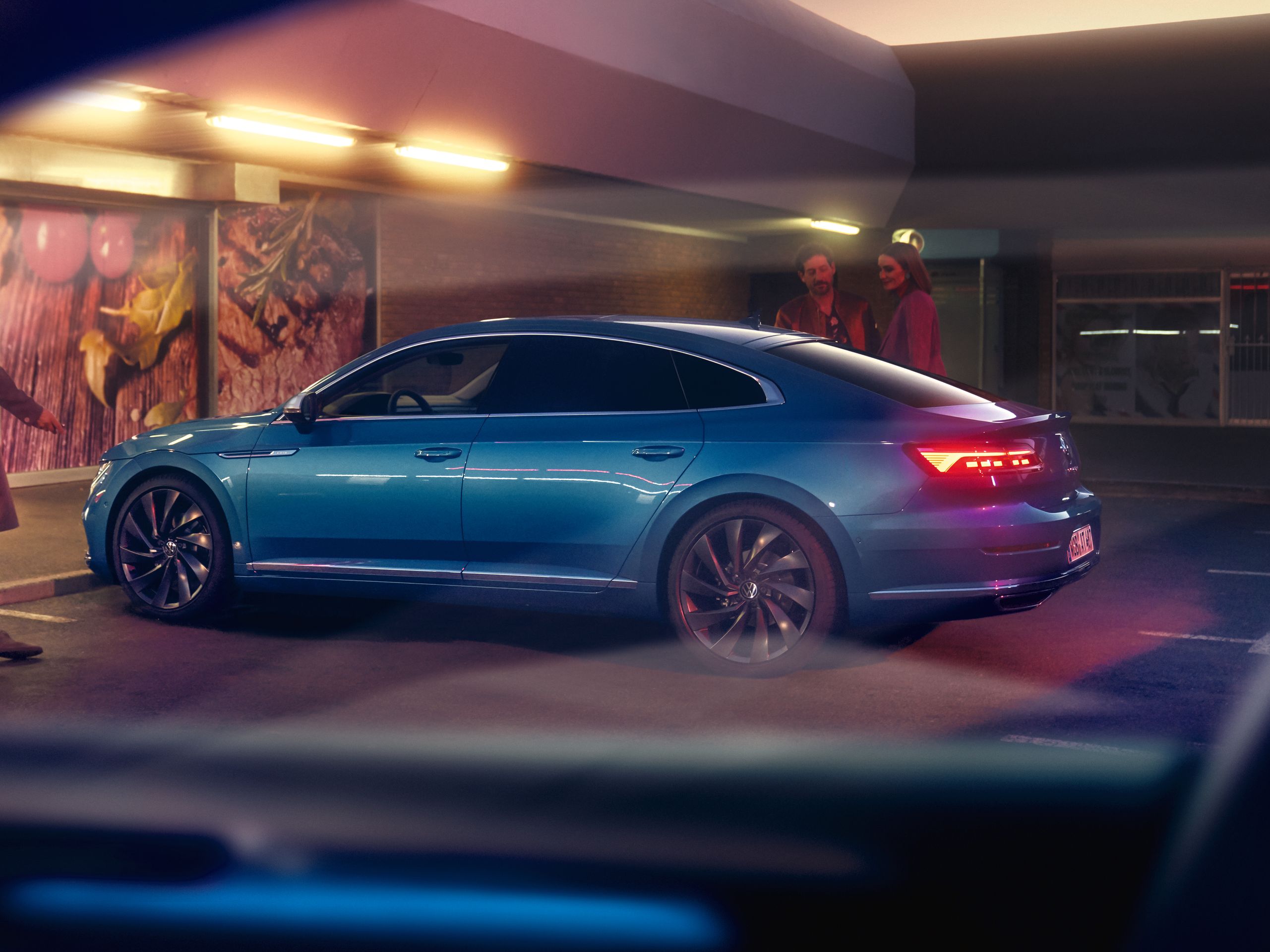 Син Volkswagen Arteon паркиран пред осветен супермаркет вечер, с хора, които разговарят наблизо