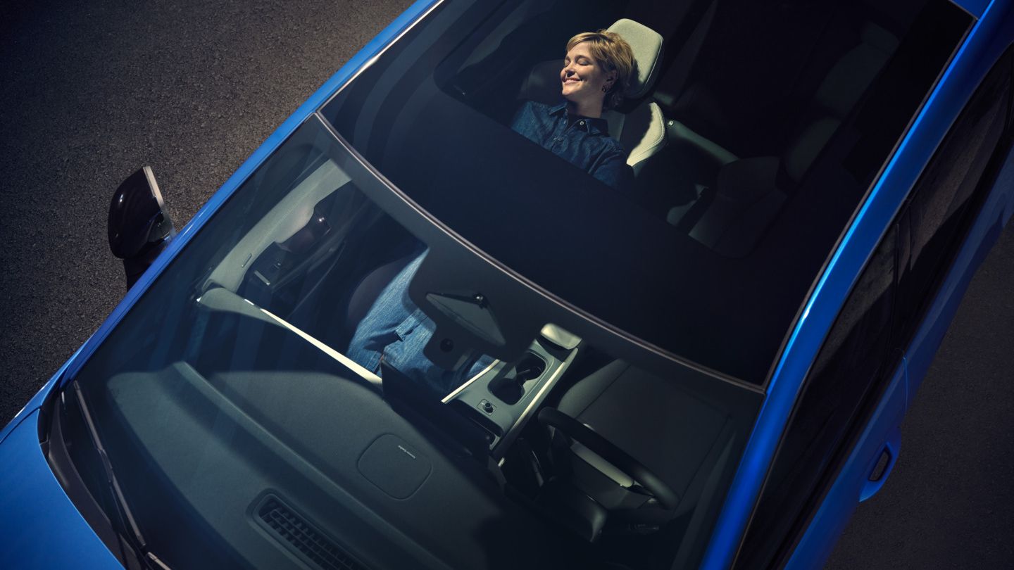 Изглед отгоре през панорамния покрив на син VW ID. Поло стил. На облегнатата седалка жена се отпуска.