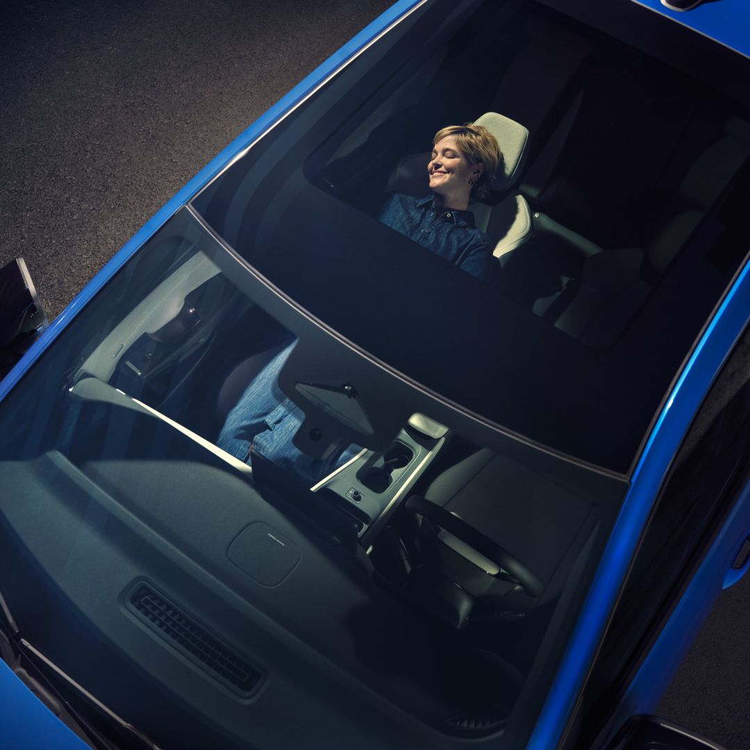 Изглед отгоре през панорамния покрив на син VW ID. Поло стил. На облегнатата седалка жена се отпуска.