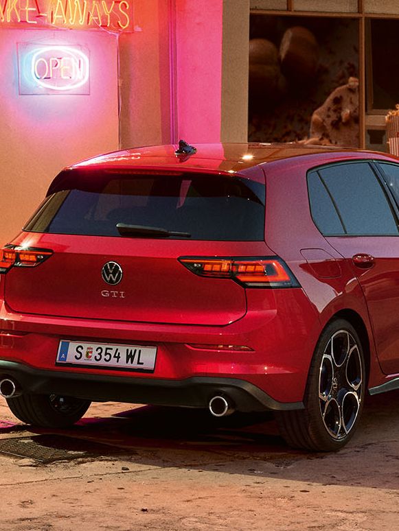 Heckansicht eines roten VW Golf GTI in abendlicher Stimmung, zwei Personen gehen neben ihm her