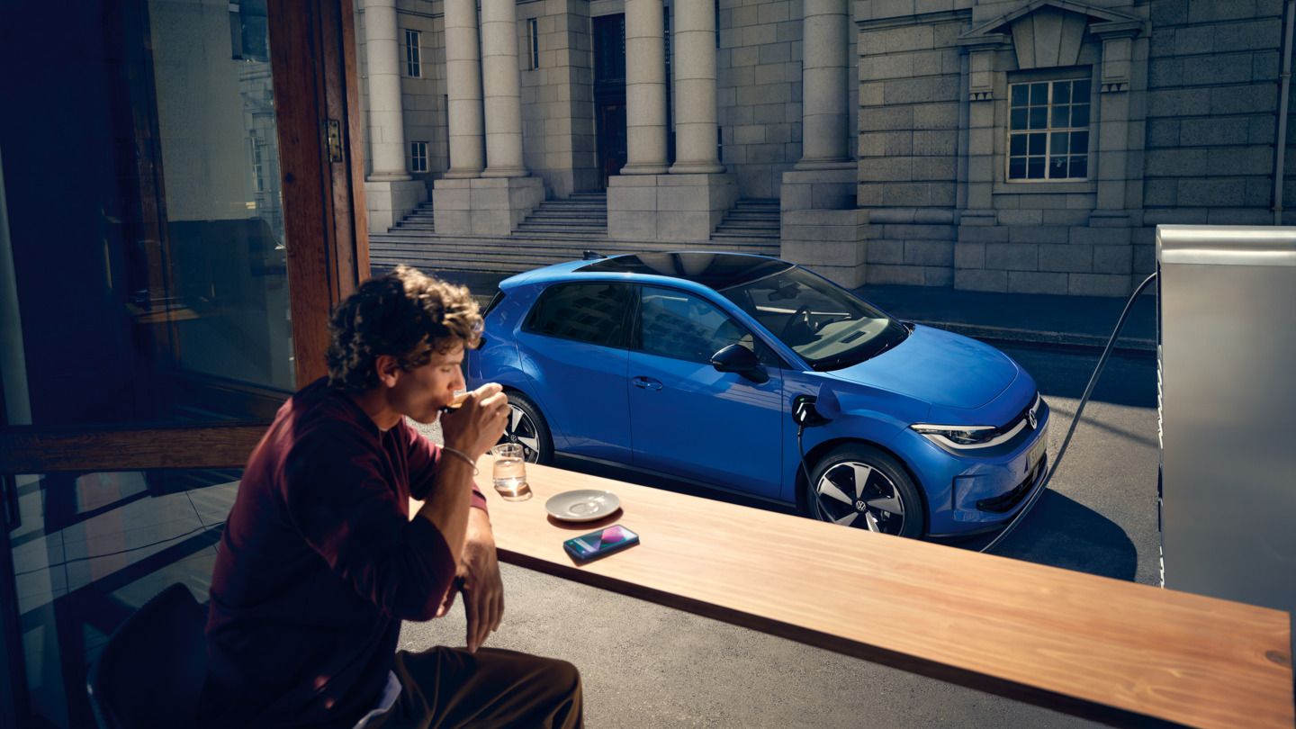 Мъж седи в кафене, през отворен прозорец се вижда синьото Volkswagen ID. Polo Style, което се зарежда на зарядна станция.