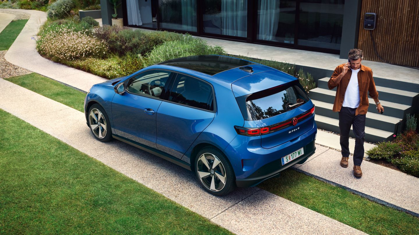 Син VW ID. Polo Style е паркиран пред зелена жилищна сграда, мъж върви зад автомобила.